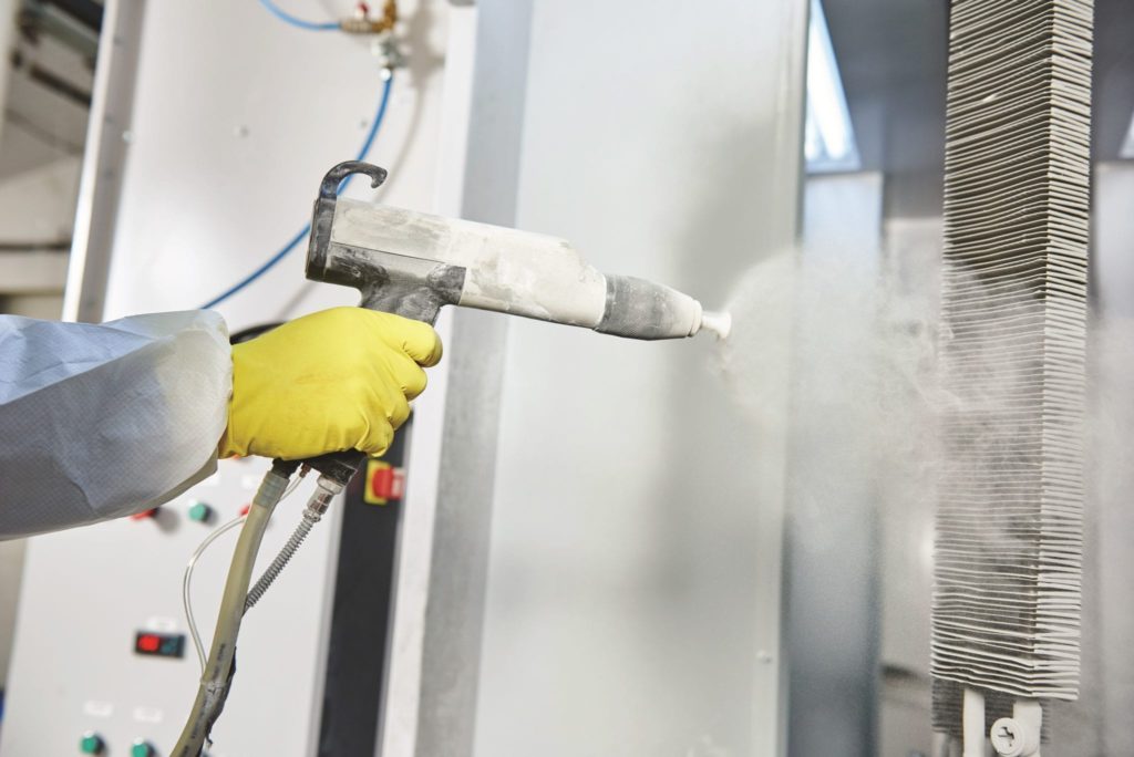 industrial metal coating. Worker man in protective suit with gas mask spraying powder to steel finished parts in painting chamber