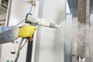 industrial metal coating. Worker man in protective suit with gas mask spraying powder to steel finished parts in painting chamber