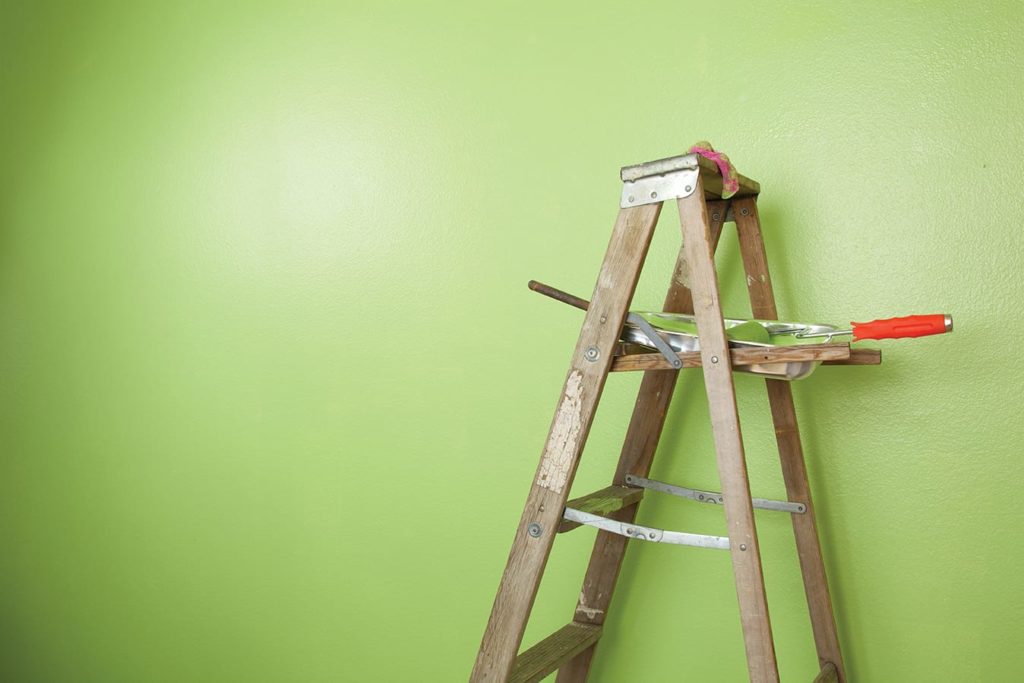 Old antique wooden ladder stands against a half painted green wall with paint roller and tray