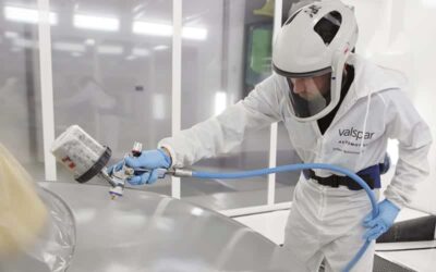 A person in white protective clothing, gloves, and mask sprays a silver surface.