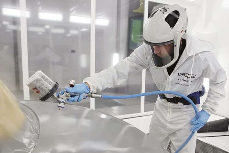 A person in white protective clothing, gloves, and mask sprays a silver surface.