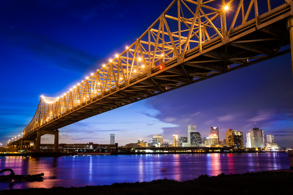 A golden bridge stretches over blue water and a dark sky. A city skyline can be seen casting jewel tones on the far shore.