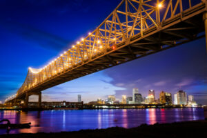 A golden bridge stretches over blue water and a dark sky. A city skyline can be seen casting jewel tones on the far shore.