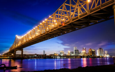A golden bridge stretches over blue water and a dark sky. A city skyline can be seen casting jewel tones on the far shore.