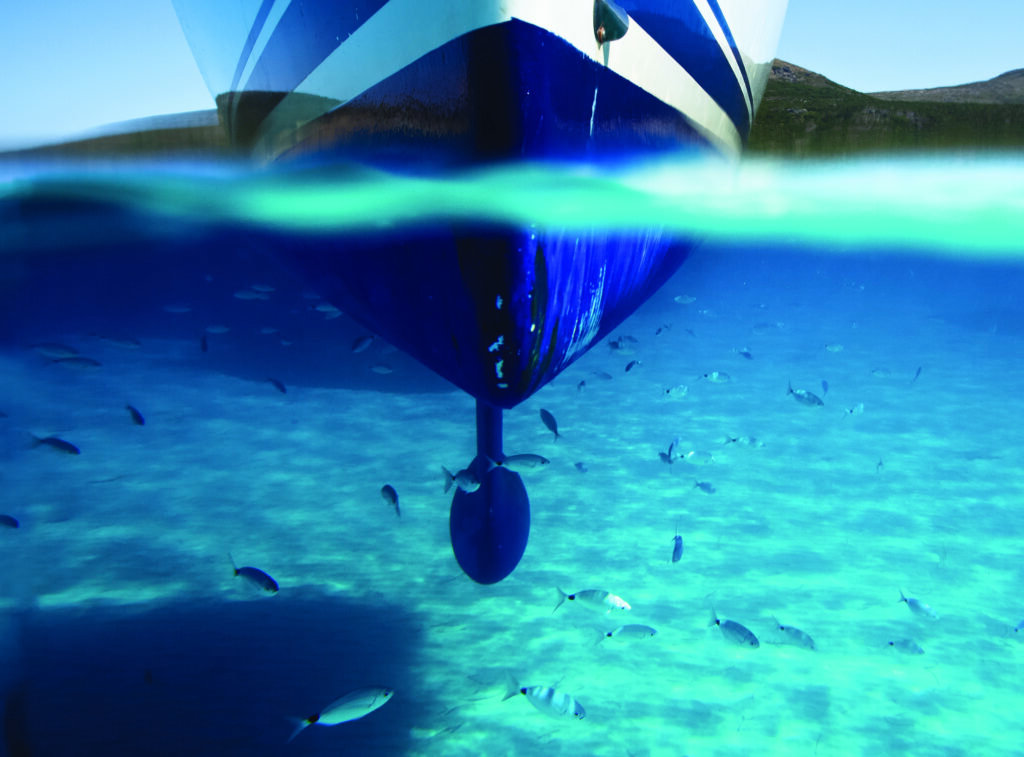 A boat seen from the front showing the hull from both above and below the water. A school of fish swims through turquoise and blue water above the sandy seafloor.