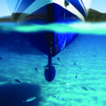 A boat seen from the front showing the hull from both above and below the water. A school of fish swims through turquoise and blue water above the sandy seafloor.